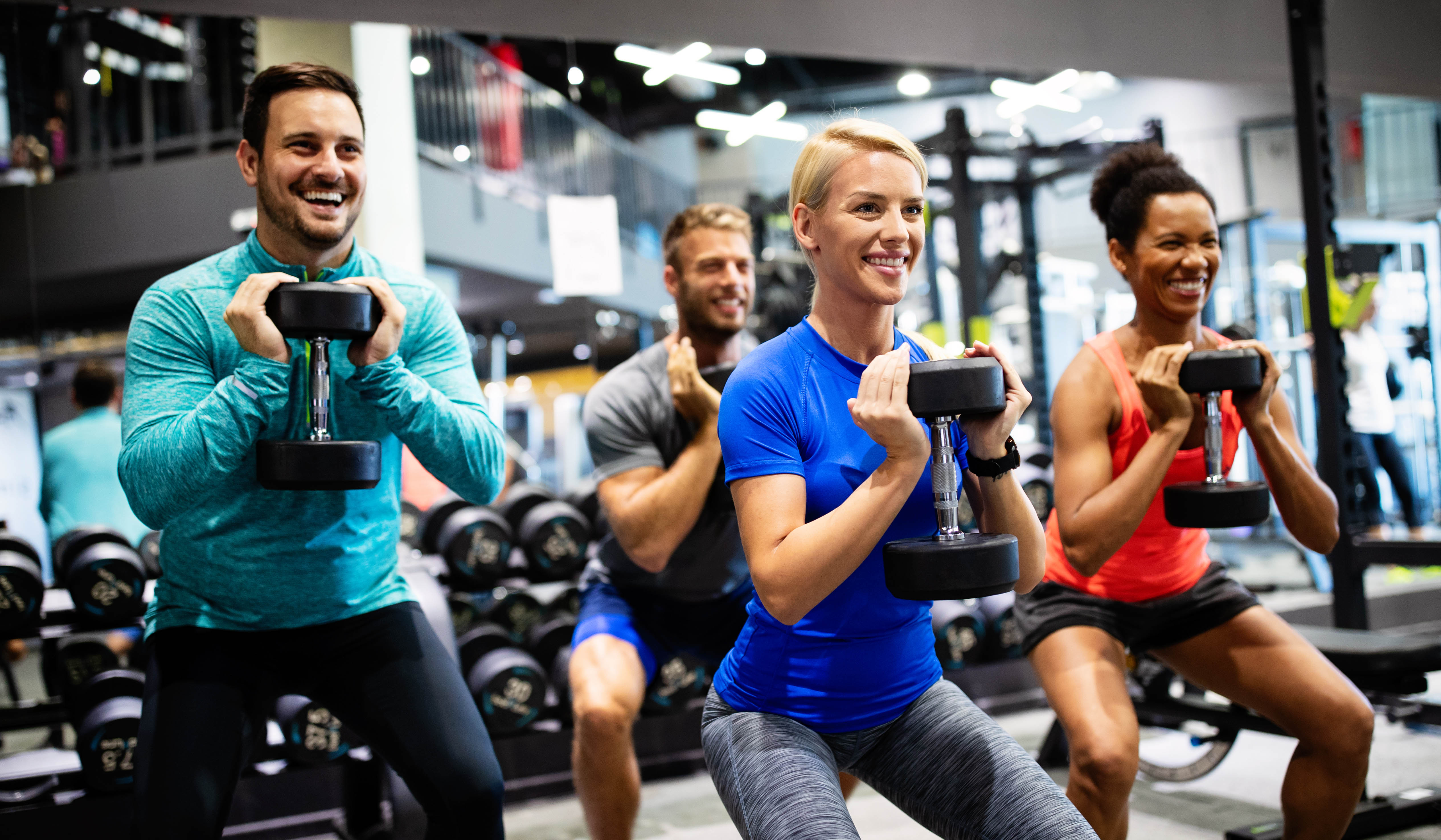 Multi-racial group of young men and women doing dumbbell squats in the gym Multi-racial group of young men and women doing dumbbell squats in the gym